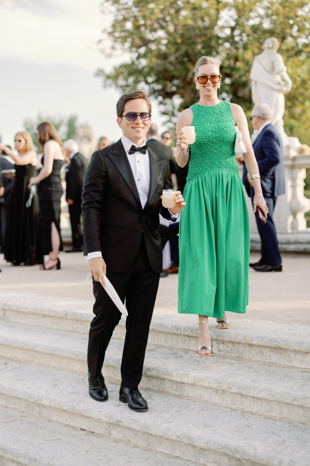 Guests in a green dress and blazer smiling with cocktails on the palace steps