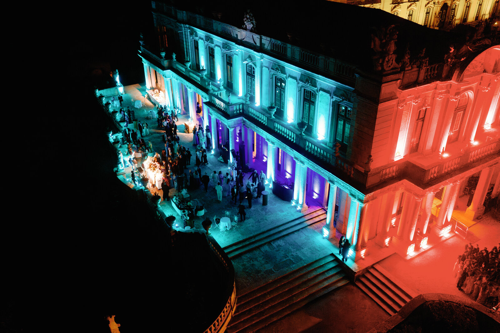 Aerial night view of the palace lit teal and red with guests gathered below