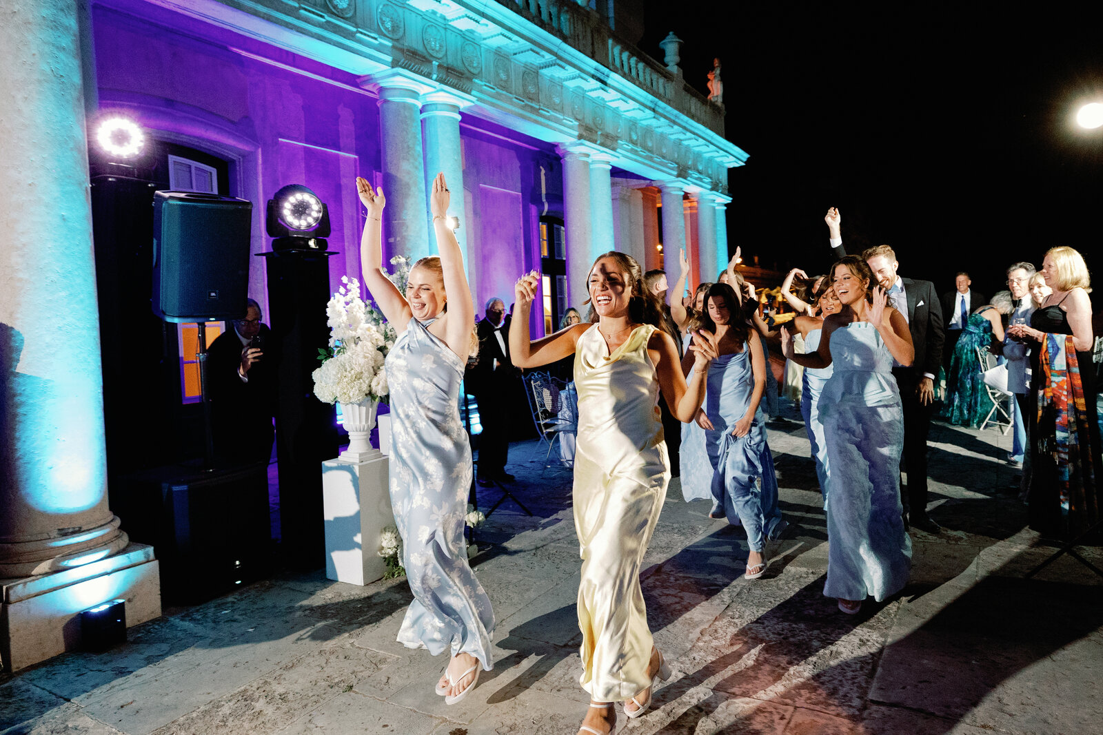 Bridesmaids dancing with arms raised in front of the lit palace