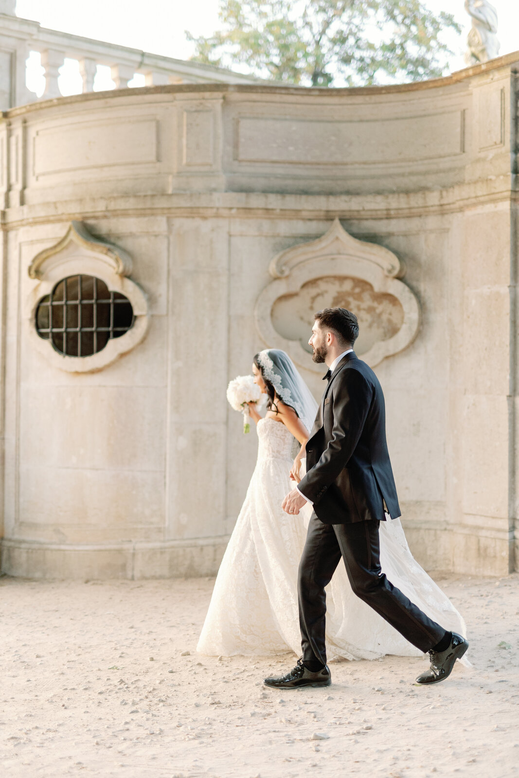 Bride and groom walking hand in hand past an ornate stone wall