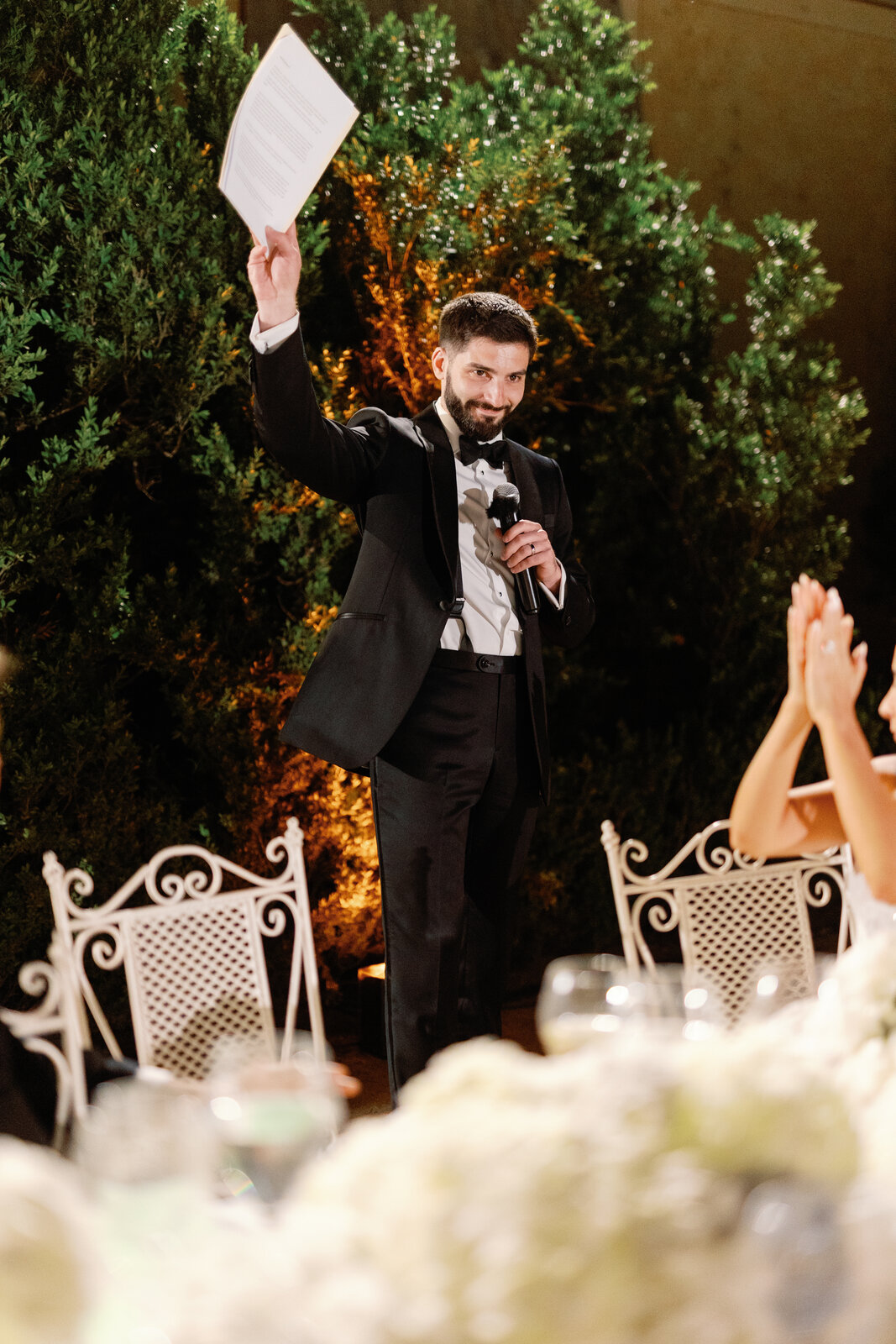 Groom giving a toast with a microphone in front of a garden hedge