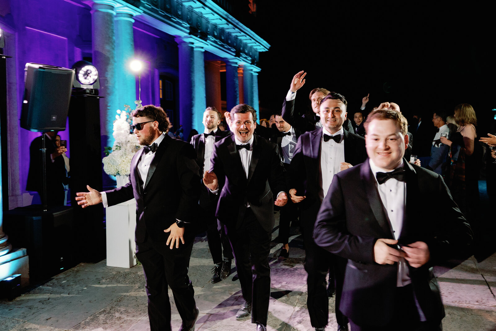 Groomsmen dancing and cheering in a line beneath the lit palace columns